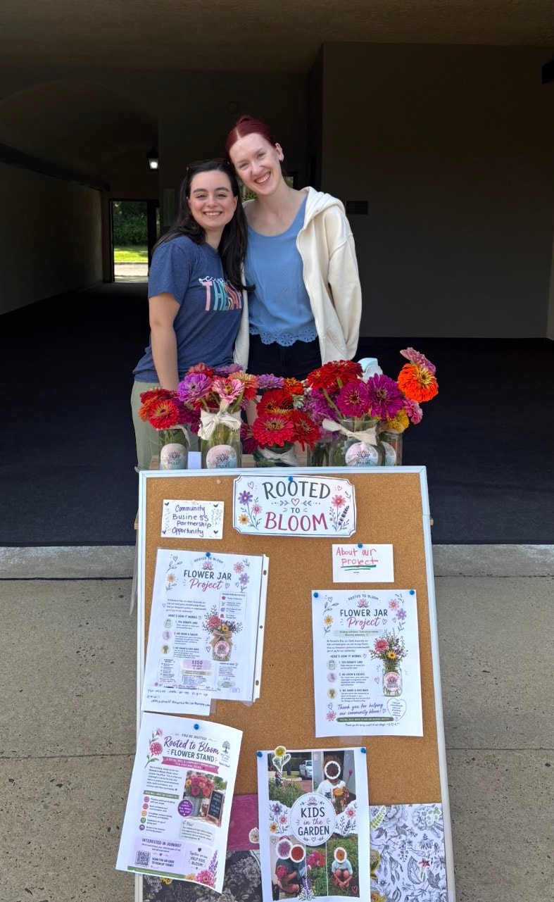 Two people smiling behind the Rooted to Bloom flower stand with corkboard display